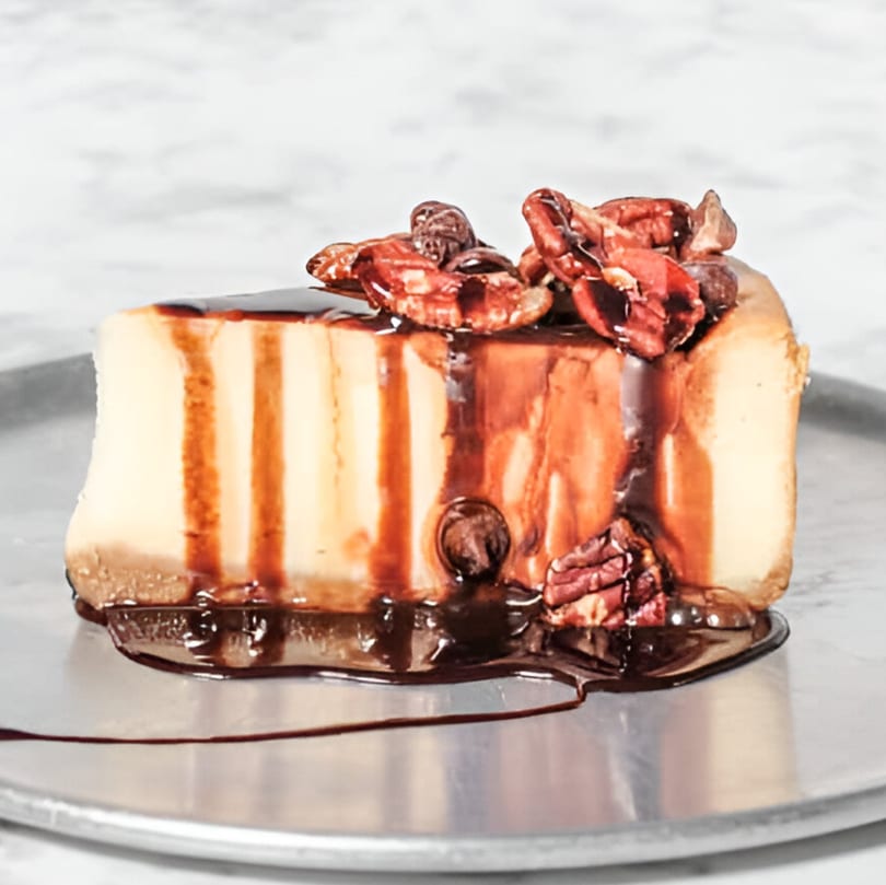 A slice of cheesecake with chocolate drizzle and pecans on a silver plate.